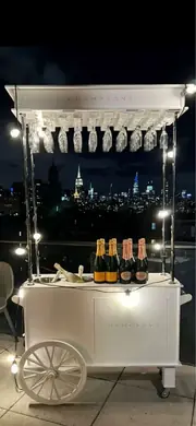 White champagne cart with string lights and bottles, city skyline behind