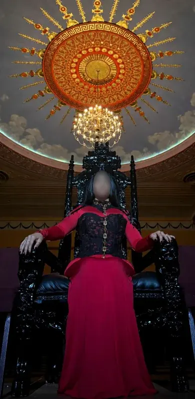 Woman in a red gown seated on the black throne under a grand chandelier